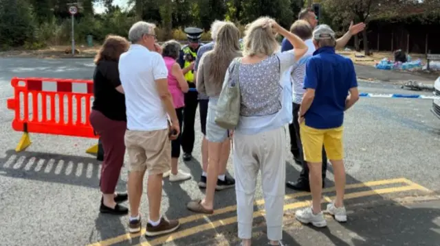 A group of people with their backs to camera stand by a police cordon, looking at someone speaking and gesticulating. One woman at the back of the group has her hand on her head.