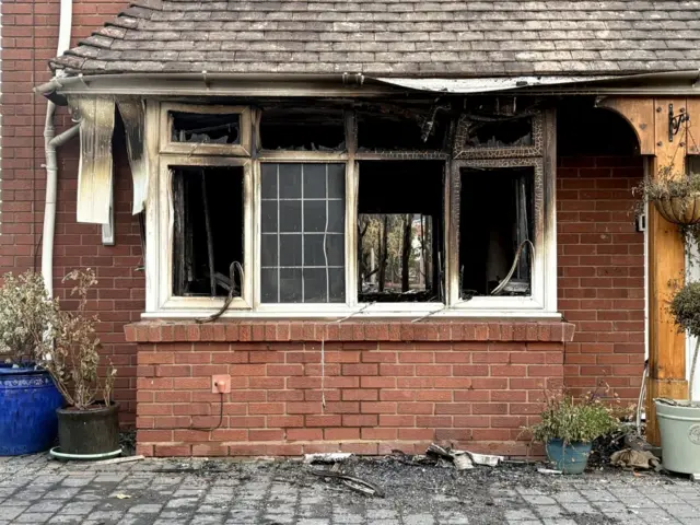 A ground floor of a red-brick home with front widows charred, warped and burned out following a fire. Plants sit in pots on a drive.