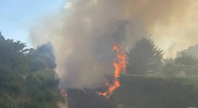 A fire on two sides of a railway embankment, with heavy smoke rising, darkening a blue sky.