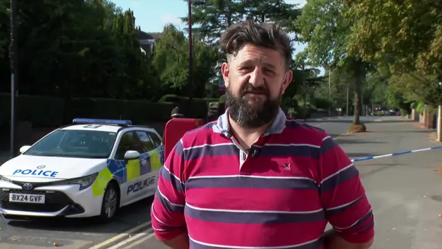 Daniel Pardoe (man in his late 40s-early 50s) wearing a rugby-style long-sleeved polo shirt in red and blue stripes, his arms crossed behind his back. He's standing in the middle of the pavement, a police cordon behind him and a police car parked to his right on a tree-lined avenue