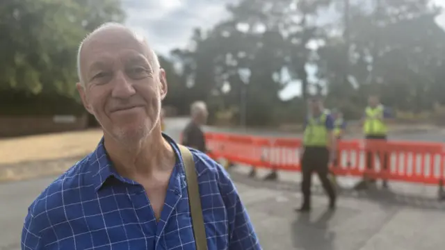 A middle aged man in a short-sleeved, blue chequered shirt poses for a picture outside a police cordon, three officers behind him in the blurred background
