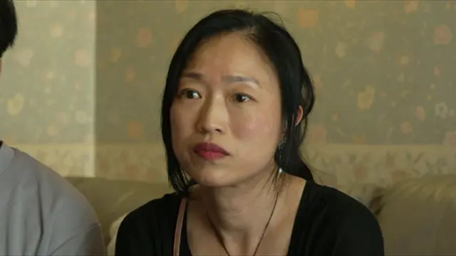 A mother sat next to her son, on a sofa, wearing a black blouse. She has black hair tied in a ponytail