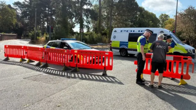 A red cordon across a road, with police car and van behind it. A man with his back to camera talks to male and female officers in high-vis yellow vests.
