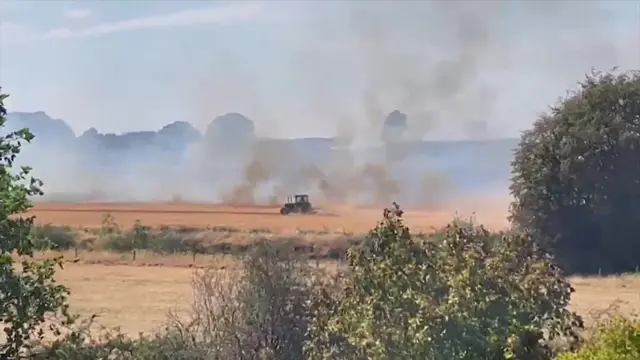 Wide shot of a field, with a tractor driving through to create a firebreak as fire approaches. Grey smoke is seen billowing up towards the sky