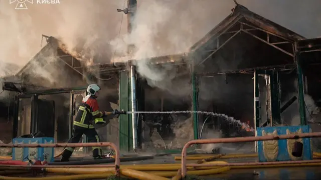 A firefighter hoses water onto a burning building, with smoke and flames rising from the roof.