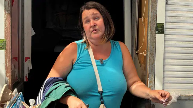 Luba leans against a wall and speaks to camera. She is wearing a blue top and is holding several jackets