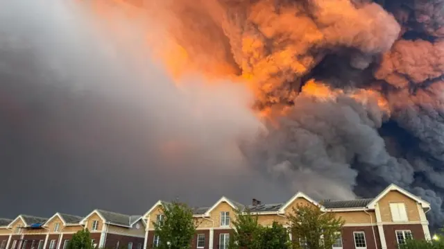 Thick smoke clouds can be seen rising behind a row of houses. Some of the smoke has turned orange