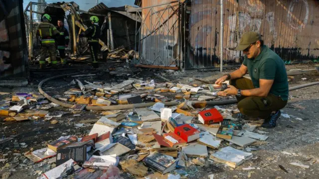 A man looks at several books on the floor that lie between debris and shattered glass