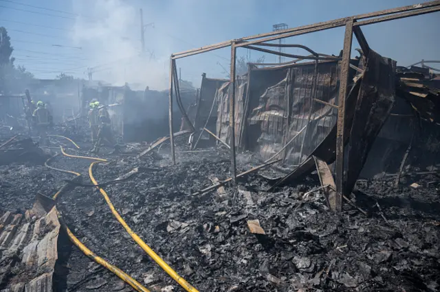 Firefighters can be seen walking through the remains of a book market. The ground is completely charred and only the outer structure of some stalls remains