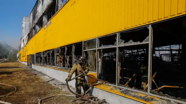 A fireman standing outside the broken windows of the market pulling a hose through the building
