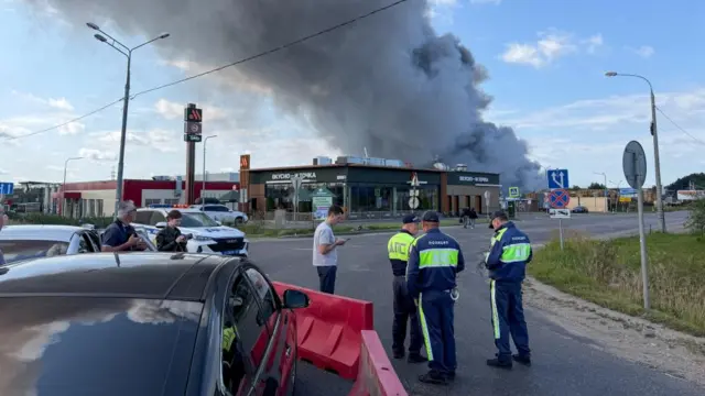 Thick smoke can be see rising from behind a retail store. Several officials can be seen standing in the road nearby