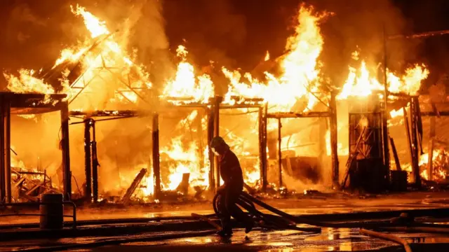 A firefighter walks past a burning building in Kyiv. Only the structure of the building remains and it has been engulfed in flames. The fire fighter is dragging a hose along