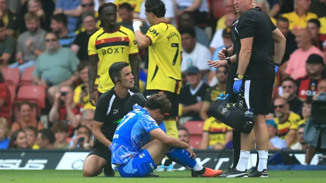 Kuryu Matsuki sits on the pitch and holds a hand to his head, as two medical staff stand in front of him talking to the referee who is also knealt on ground