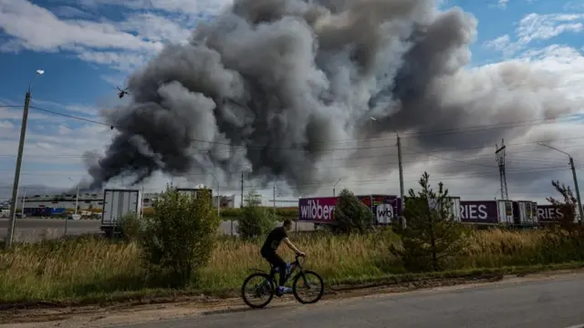 A person rides a bicycle along a road near a burning Wildberries facility. Large clouds of thick, dark smoke can be seen in the background