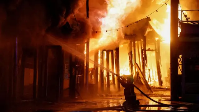 A firefighter sprays water from a hose towards flames. Market stalls can be seen burning in large flames in the background