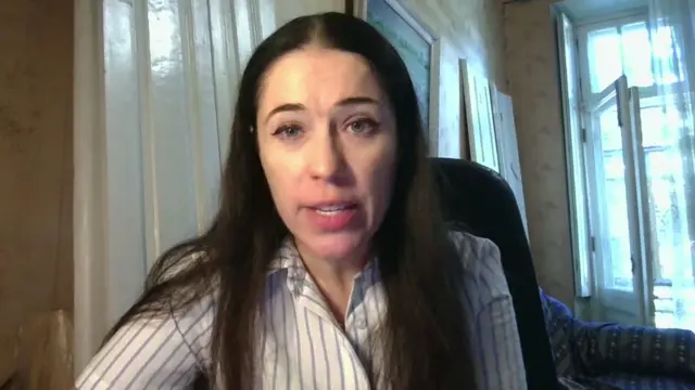 Hanna Shelest, a woman in her 30s with long brown hair, wearing a striped, long-sleeved shirt, speaks to the computer camera during a remove interviews. She's sitting in an office chair inside what appears to be a room in a residential building