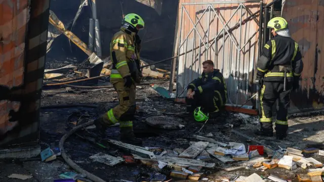 Two firefighters stand and one sits down next to damaged books on the floor, Debris is spread around them including pieces of wood and the earth has been charred and appears black