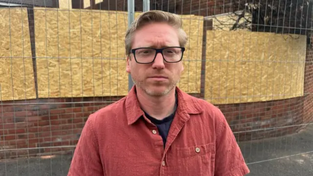 A man with blonde hair, glasses and a red shirt standing in front of metal fences and a boarded up building