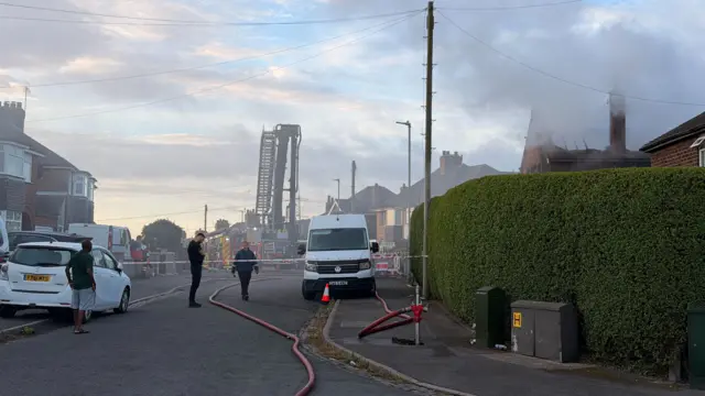 A cordon on a residential road as firefighters tackle a fire. A fire engine and aerial-platform can be seen in the distance. Red and white tape is stretched across the road in front of us, while people stand or walk around.