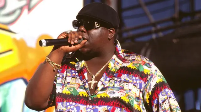 Biggie wearing a patterned colorful shirt and big gold cross necklace performing into a microphone