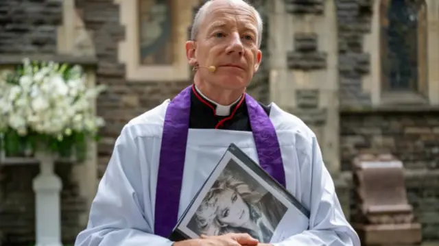 A reverend canon is wearing ceremonial attir, and is holding a picture of Bonnie Tyler in his hands