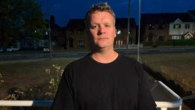A man with short hair and a black t-shirt, stands in front of a white railing next to a residential street at night, with a solemn look on his face.