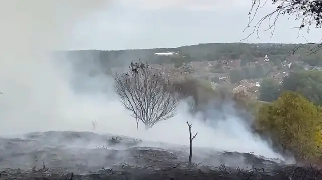 A view from a hillside charred by a fire with smoke rising from the ground. A village or town is visible in the distance.
