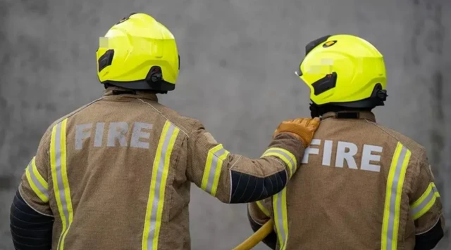 Two firefighters facing away from the camera with beige protective coats and yellow helmets