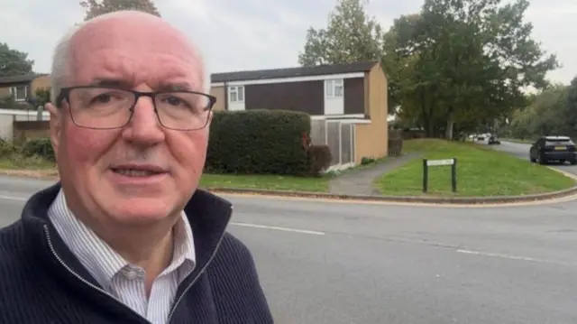 A man with thin grey hair, glasses and a blue fleece in a street with houses on the far side