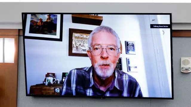 a TV screen in a courtroom showing the face of Brent Becker via Zoom
