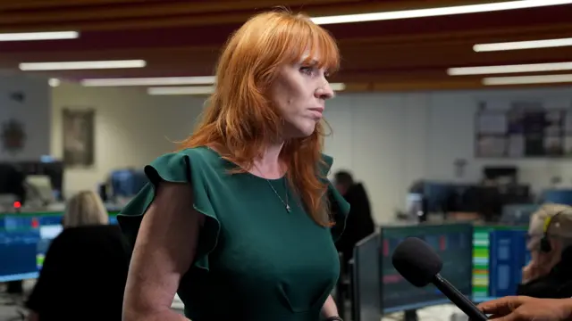 Angela Rayner has long ginger hair and is wearing a forest green dress. She is standing in a fire control room with people at computers with headsets on. Someone is holding a black microphone out to her