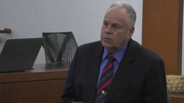 An older man with short, white hair wears a navy suit and a tie while sitting in the witness stand in court.