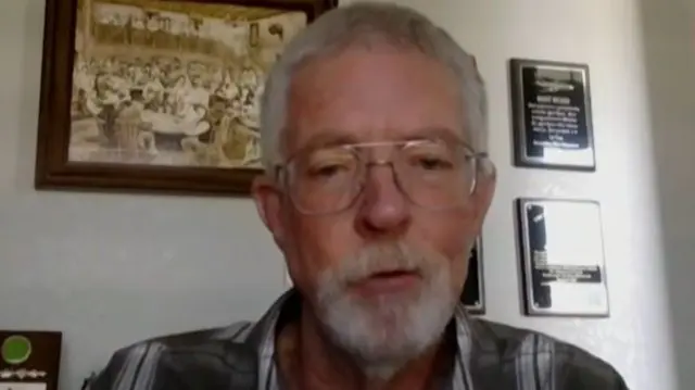 A man with grey hair and a grey beard with wire-framed glasses sits in a room with various framed plaques and images behind him