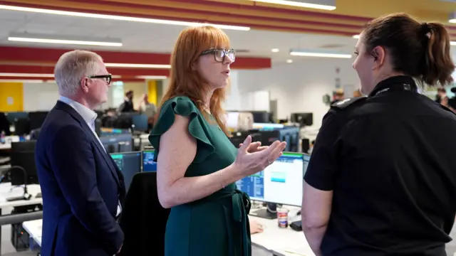 Angela Rayner, wearing a green dress, speaking to a woman in a fire control room. Richard Parker, West Midlands mayor, is standing on the other side of her.