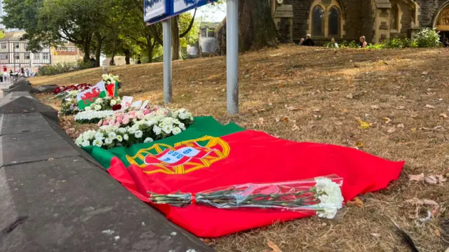 A large Portuguese flag is laid on the ground among floral tributes