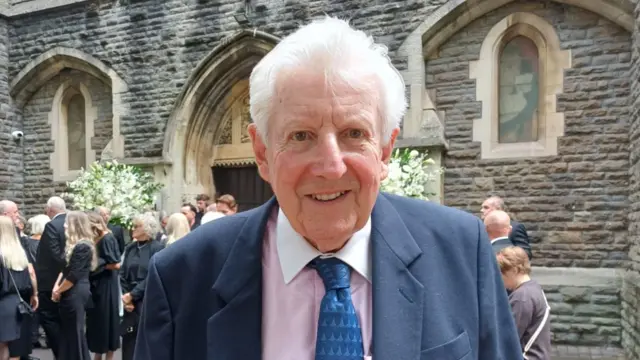 A man looking at the camera. He has white hair, a blue jacket and tie and light pink shirt.