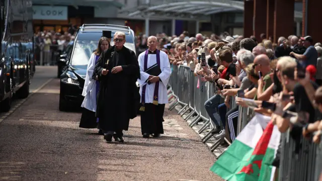 Three people are walking in front of a hearse on a street, and to the right are many people stood behind a barrier watching on