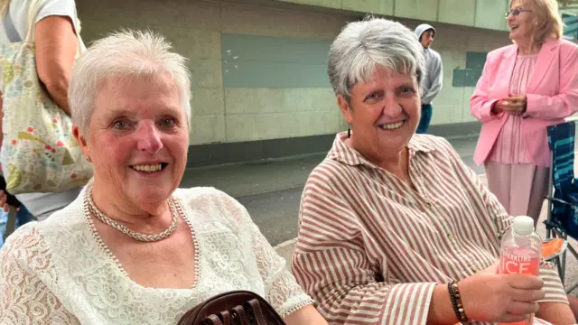 Two women are sat down in chairs, looking at the camera and smiling