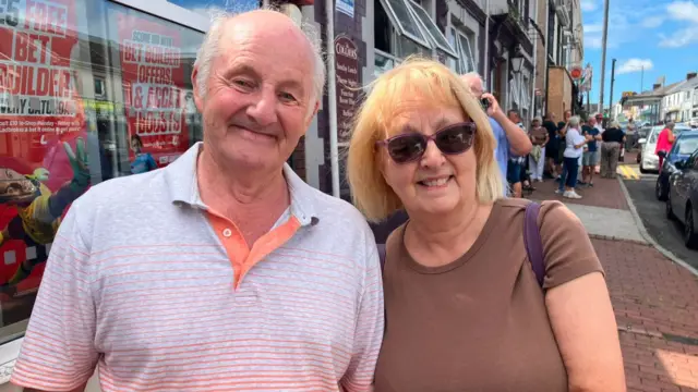 A man is wearing a grey polo shirt, smiling at the camera and is stood next to a woman who is also smiling