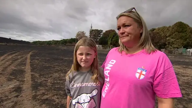A woman with long blonde hair, sunglasses on her head and wearing a pink England shirt, stands next to a girl with long blonde hair and a grey shirt. They are both on a field that has been charred by fire.