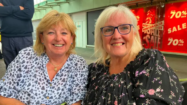 two women sit in deck chairs on a pavement with a shop and sale signs in the background. the one on the right in a black blouse wiith flowers, neck length blonde hair and black glasses has her am around the second woman on the right who has similar hair and a white blouse with blue flowers. both are smiling