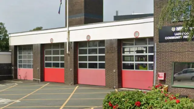 Four red doors on the front of a fire station with a green bush in the foreground