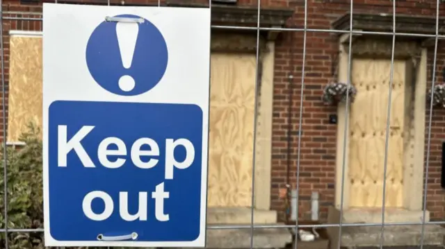 A metal fence with a blue keep out sign in front of a boarded up building