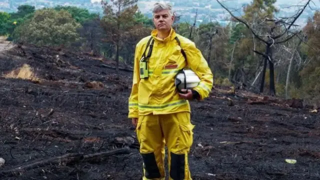 A man with grey hair, wearing a bright yellow firefighter's outfit and with a white helmet under his arm, stands on charred grassland following a wildfire.