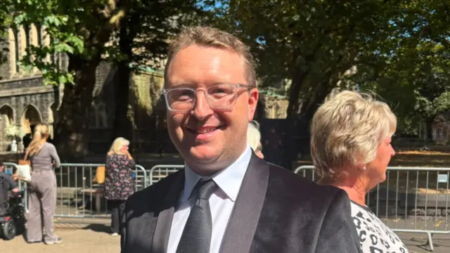 a man in clear rimmed glasses and a black suit smiles for the camera on the street in swansea on a sunny day