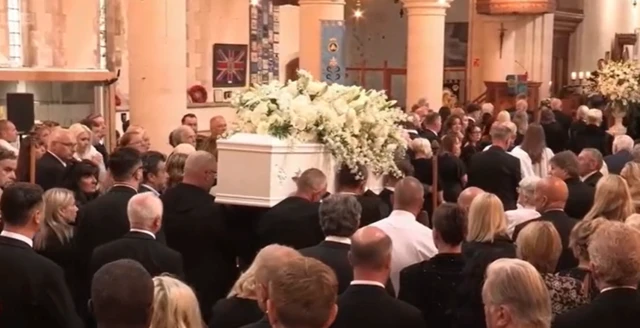 People are stood inside a church watching a large white coffing be carried through