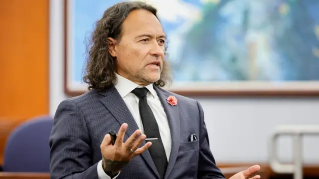 A man with long hair and a dark suit standing in a court room.