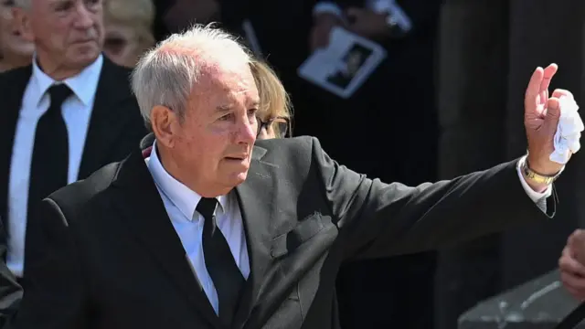 A man in a black suit waving. He is visibly upset.