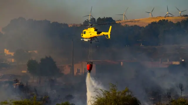 A yellow helicopter carrying a red container which is releasing water over burning trees