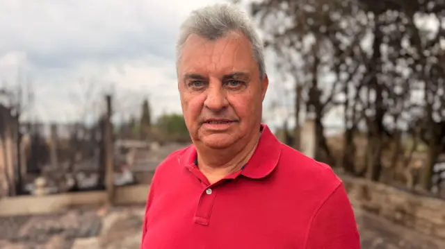 A man with a red shirt stands in the charred remains of his back garden.